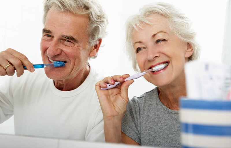 An older couple smiling while brushing their teeth