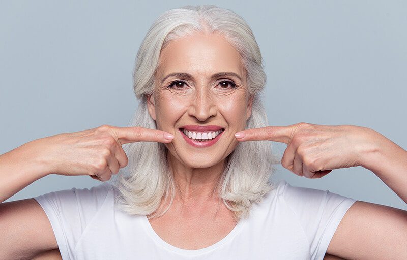 A white haired woman pointing to her mouth with both hands