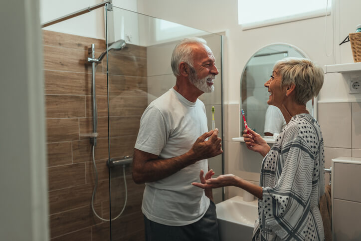 An older couple smiling at each other while brushing their teeth