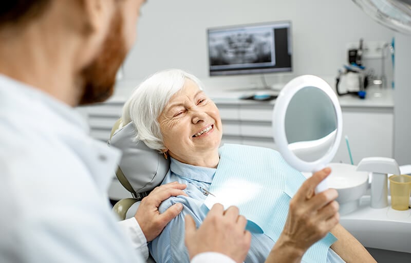 An older woman looks at her teeth in a hand mirror