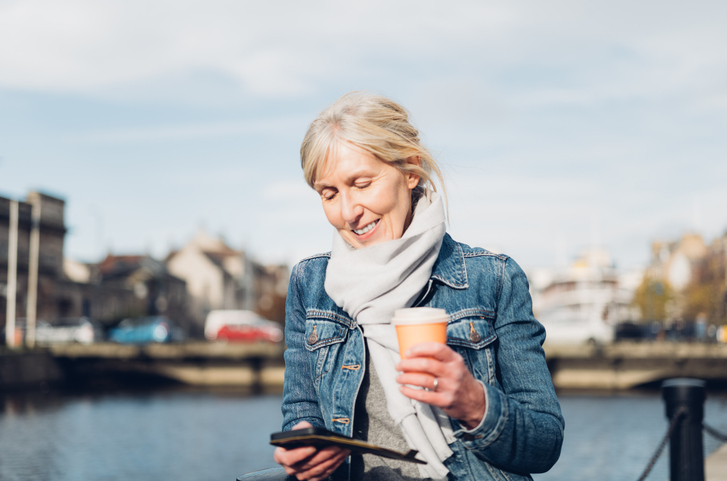 Mature woman with smart phone and coffee beside the riverfront at Leith in Edinburgh, Scotland.