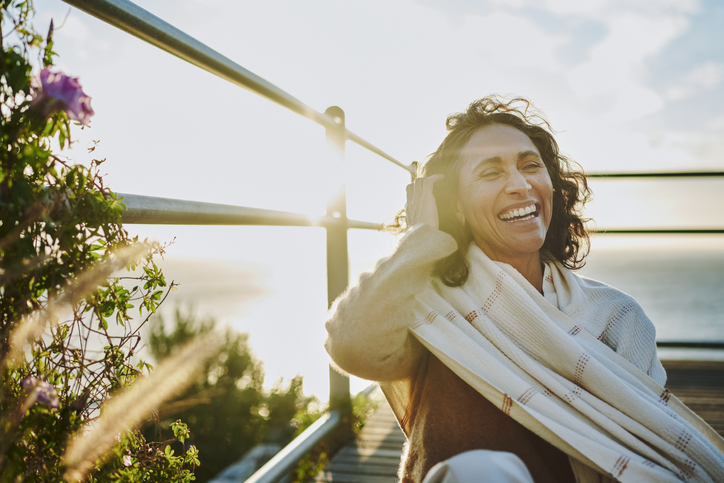 Mature woman wearing a pashmina laughing while sitting at a scenic viewpoint by the ocean at sunset