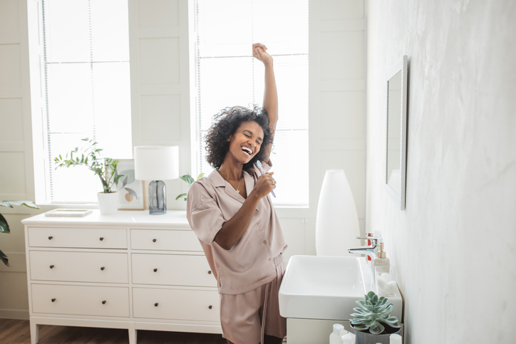 Young woman in pajamas looking in mirror and brush teeth with toothbrush in home bathroom.