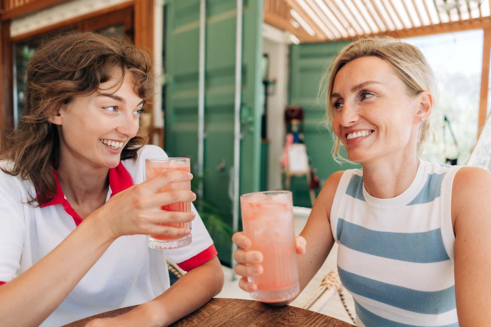 Two friends laugh and toast cold drinks while enjoying outdoor cafe on a sunny day.