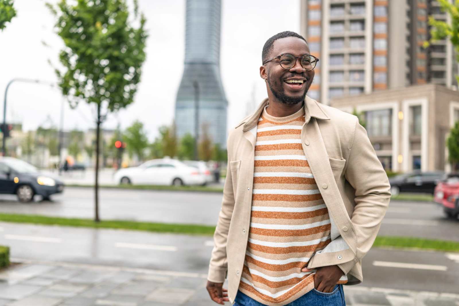 A man walks joyfully along the city street. He wears a casual outfit and glasses. Cars are parked nearby, and the buildings surround him.