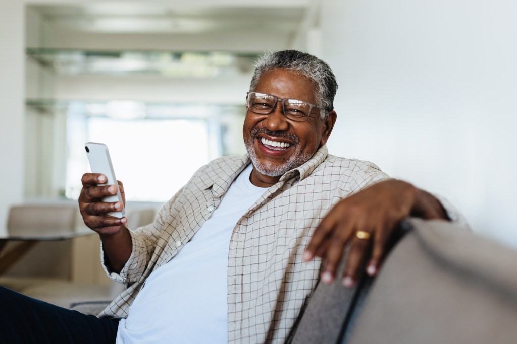 Cheerful man enjoying modern technology at home, sitting comfortably on the couch and smiling joyfully while holding a smartphone