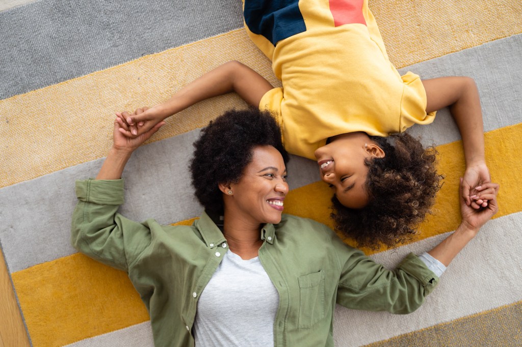 High angle view of beautiful happy African American mother and daughter holding hands while lying on the floor in the living room looking at each other and smiling.