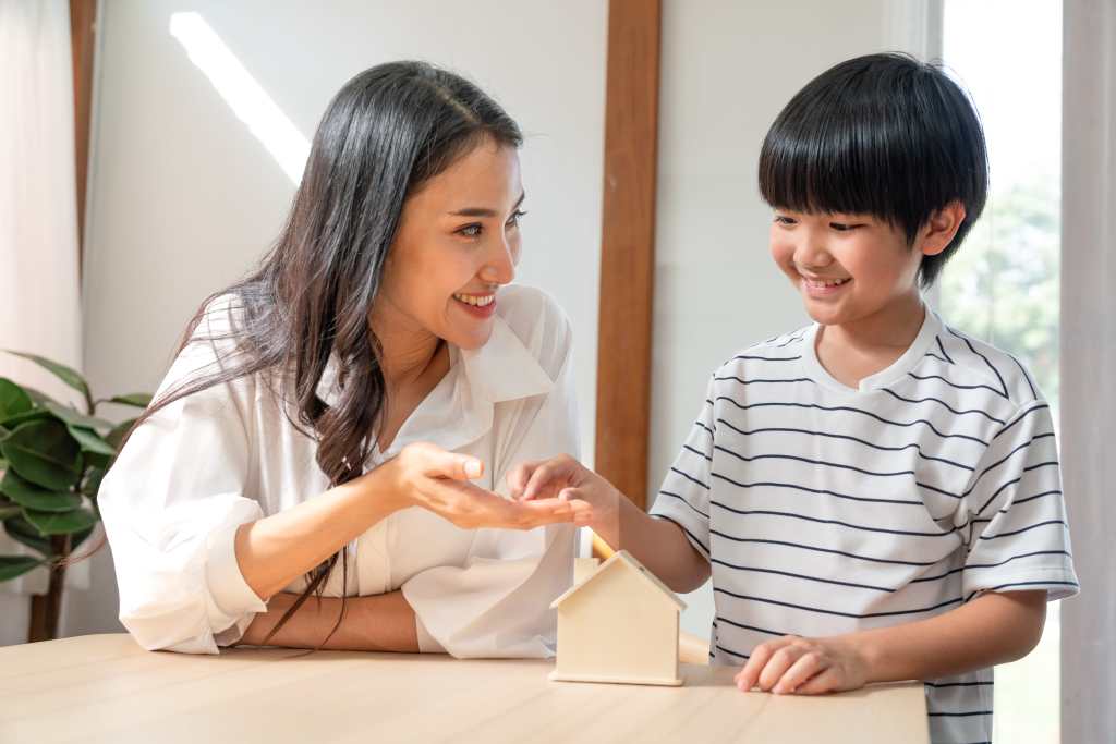 Asian mother teaches son to save money put coins into small wooden house model bank. Happy family