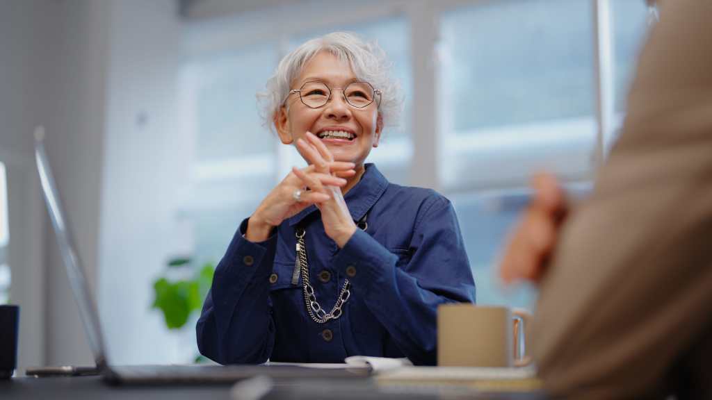 A professional older businesswoman in glasses and a navy blouse offers financial guidance to a senior businessman in a beige suit. They analyze financial charts displayed on a laptop screen, discussing key investment strategies. The collaborative setting in a bright, modern workspace highlights expertise, digital finance, and strategic decision-making.