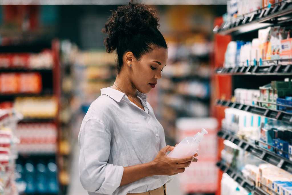 A young woman examines a product in a supermarket aisle, showcasing everyday shopping activities. The modern setting emphasizes consumerism and choice.