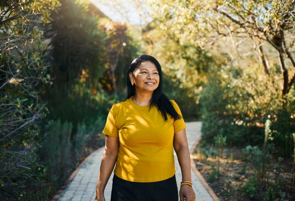 A cheerful woman with long black hair and a yellow shirt walks along a sunlit garden pathway surrounded by lush greenery and trees.