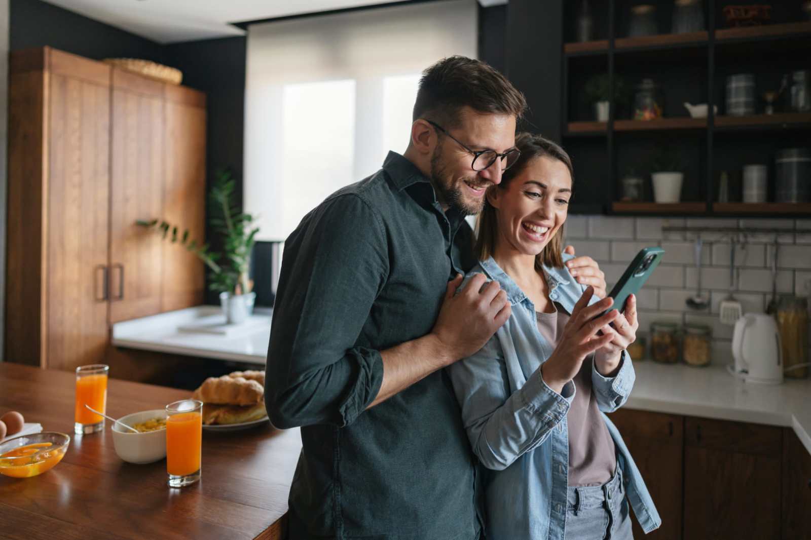 Young couple sharing a joyful moment while browsing their smartphone during breakfast in the kitchen, smiling and enjoying their time together over delicious food and drinks